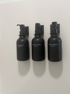 Three black bottles labeled 'SHAMPOO,' 'CONDITIONER,' and 'BODY WASH' against a white wall.