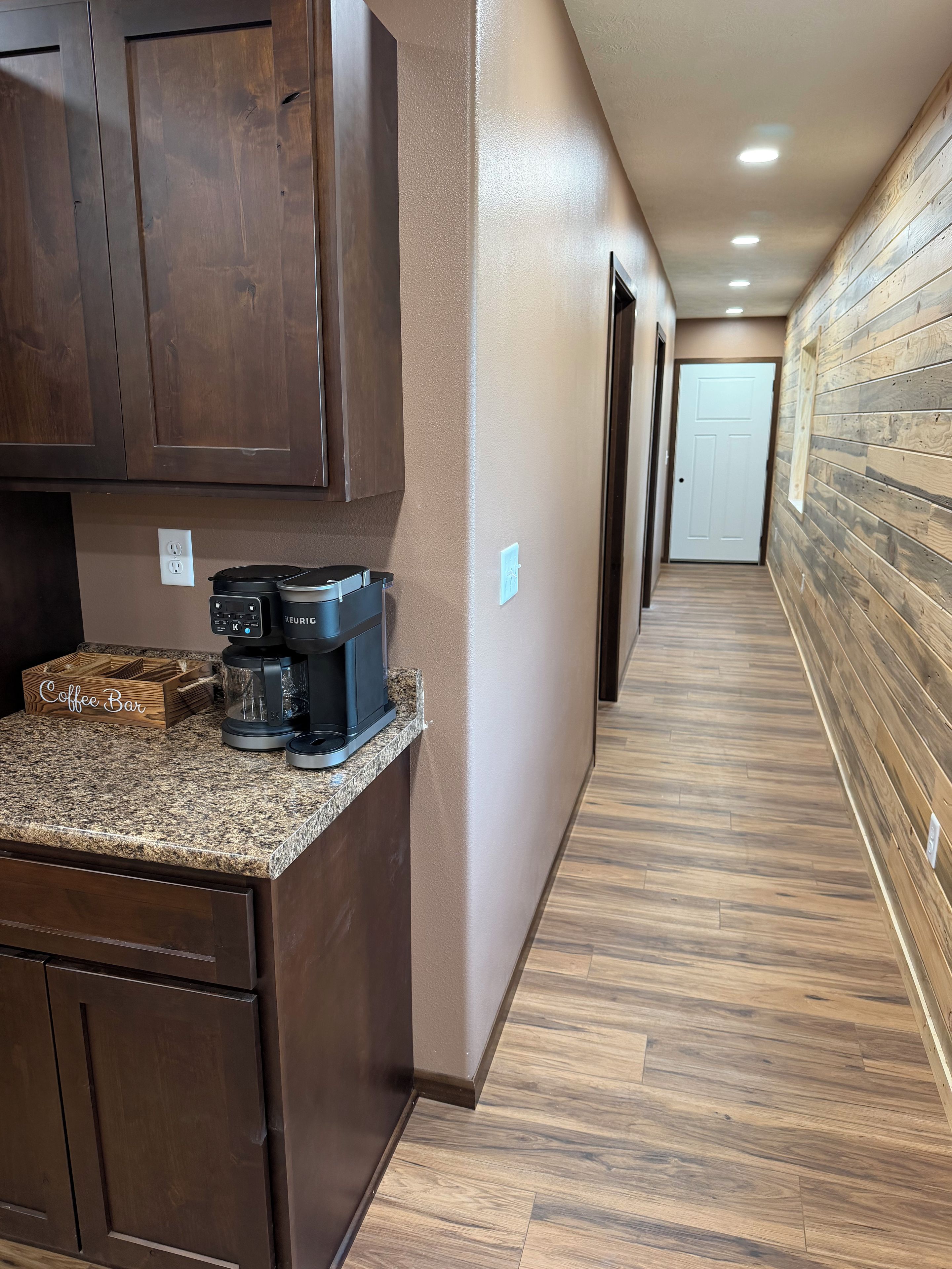 Un corridoio con pavimento in legno, un'area con un angolo caffetteria e una macchina Keurig, e una porta in fondo.
