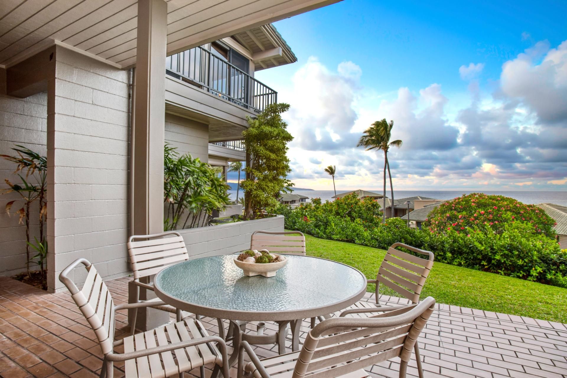 A patio with a round glass table and four chairs, overlooking a lush garden and a view of the ocean.
