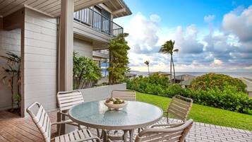 A patio with a round glass table and four chairs, overlooking a lush garden and a view of the ocean.