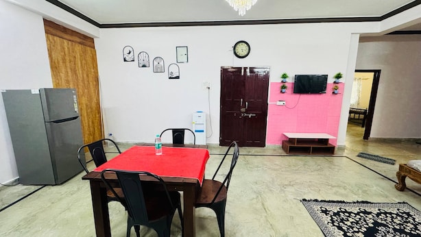 Espaço de refeições com mesa, cadeiras, geladeira, bebedouro e relógio na parede.