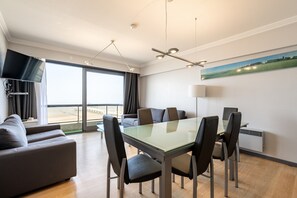 Ein modernes Hotelzimmer mit Esstisch, Stühlen, einer Couch, einem Flachbildfernseher und einem großen Fenster mit Blick auf den Strand.