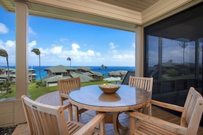 Un área de comedor al aire libre con techo, con una mesa de vidrio y sillas de madera, con vista a un resort con varias edificaciones y vista al océano.