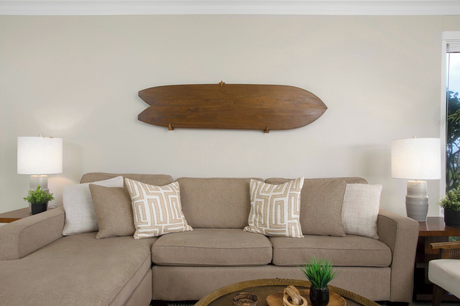 A living room with a beige sectional sofa, two patterned throw pillows, a wooden wall art piece, and two bedside lamps.