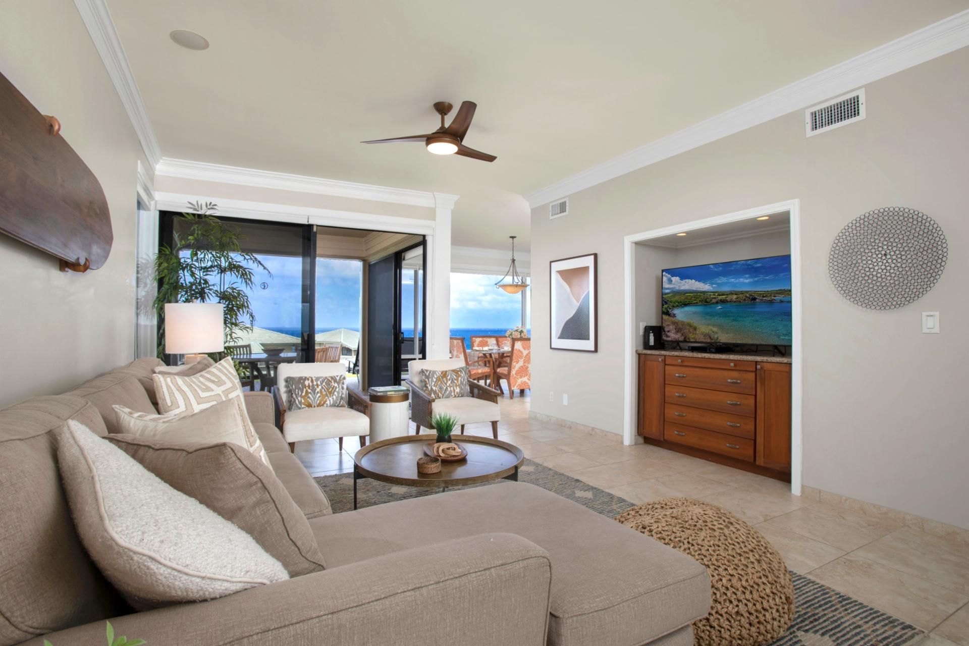 A spacious living room with a large sofa, a coffee table, and a TV in the corner. There is a dining area with chairs and a table, and a ceiling fan.