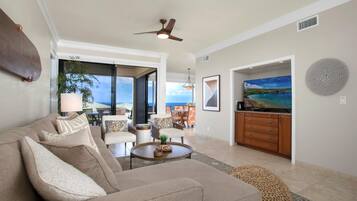 A spacious living room with a large sofa, a coffee table, and a TV in the corner. There is a dining area with chairs and a table, and a ceiling fan.