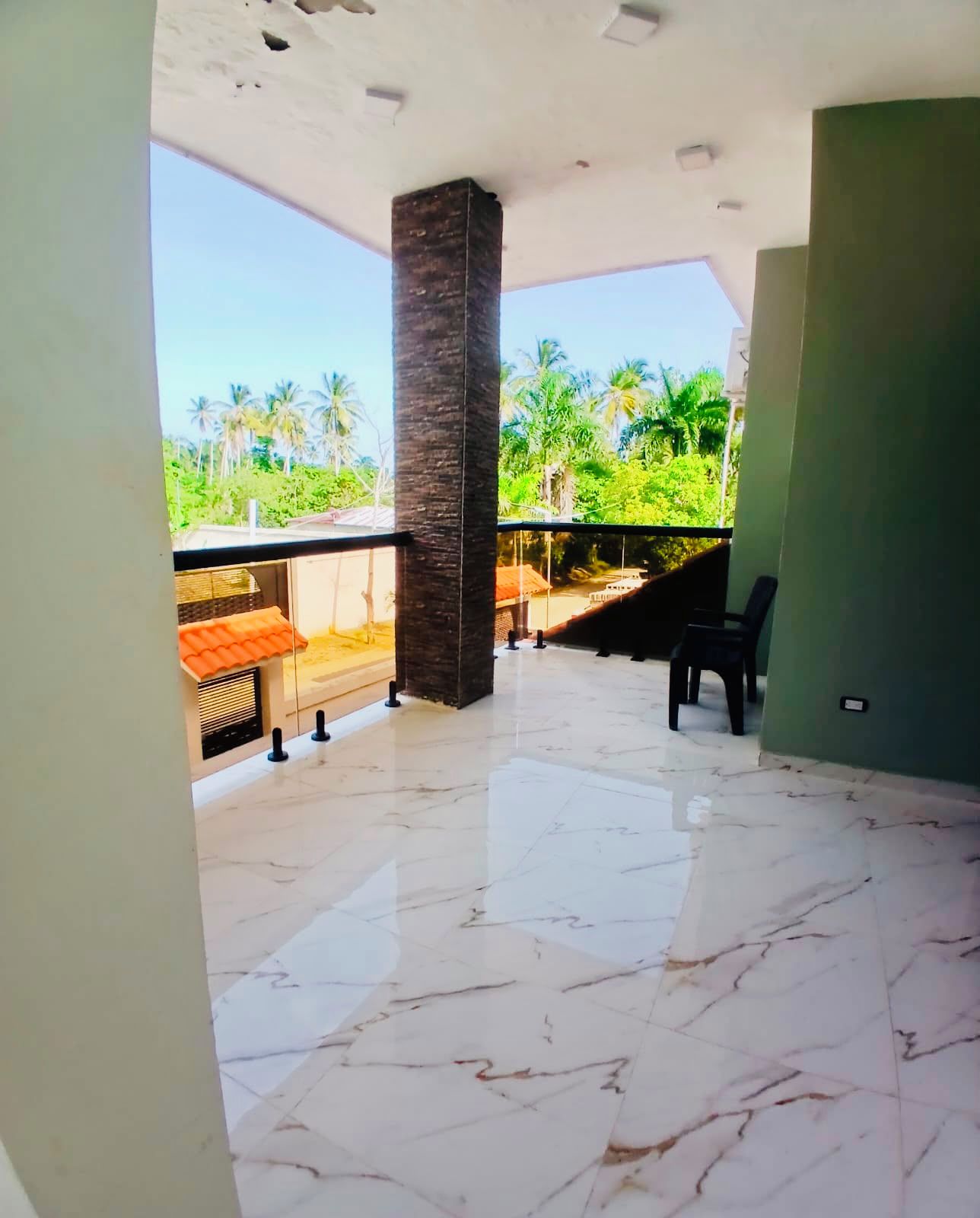 A room with a marble floor, a large pillar, a view of palm trees, and a balcony with a chair.