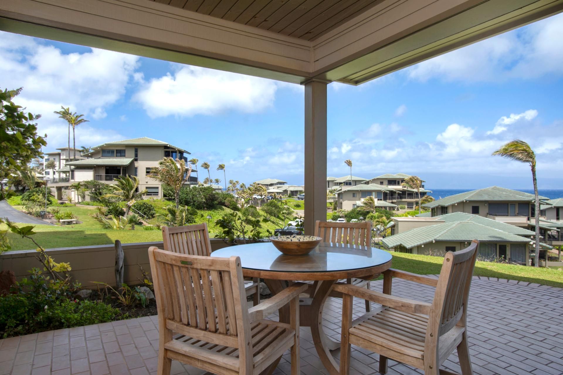 A covered outdoor seating area with a round table and wooden chairs, overlooking a residential area with houses and a view of the ocean.