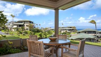 A covered outdoor seating area with a round table and wooden chairs, overlooking a residential area with houses and a view of the ocean.