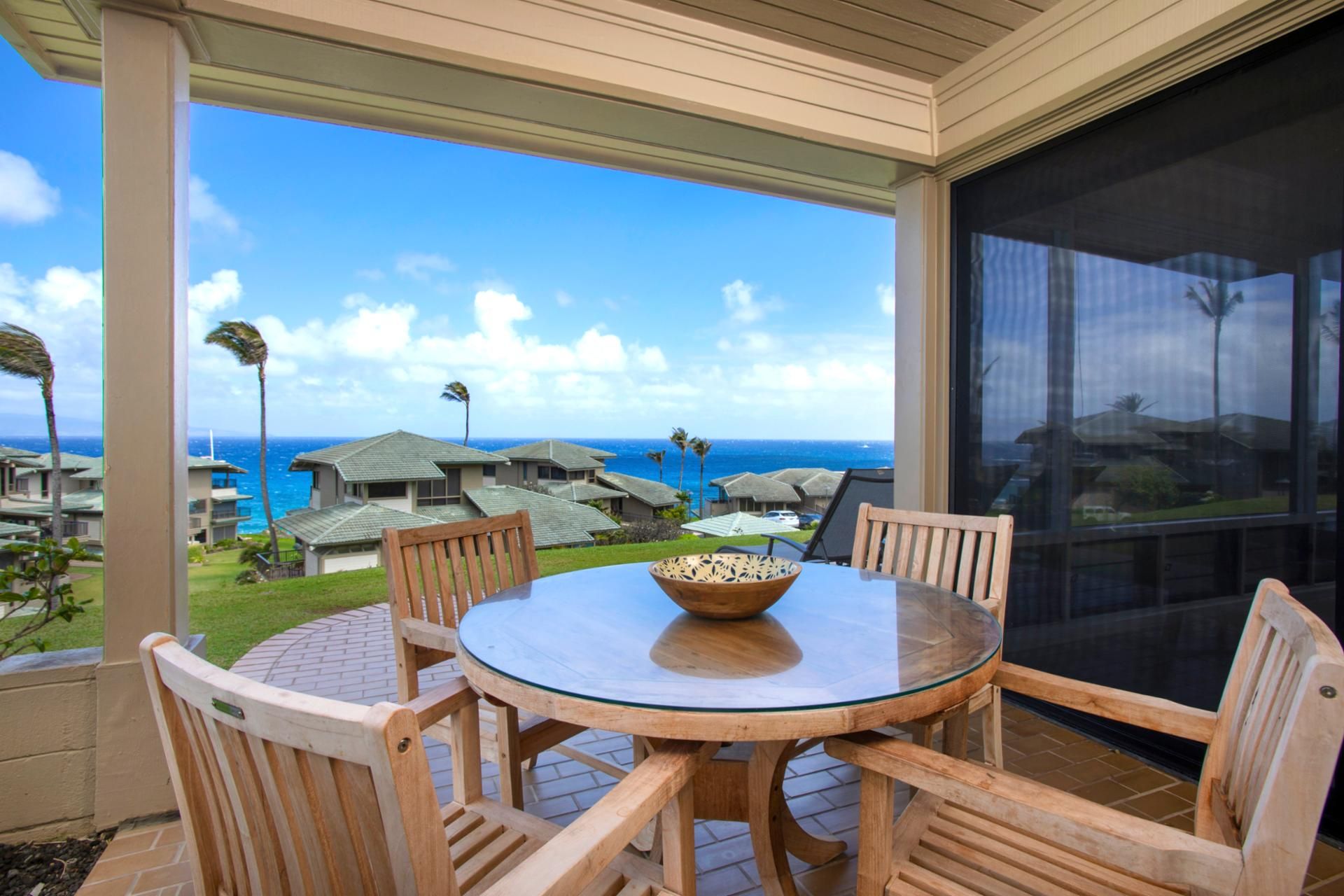 Un espace extérieur abrité, doté d’une table en verre et de chaises en bois, offrant une vue sur la côte, avec des maisons et l’océan.
