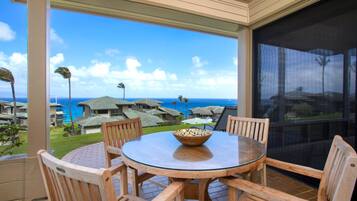 Un espace extérieur abrité, doté d’une table en verre et de chaises en bois, offrant une vue sur la côte, avec des maisons et l’océan.