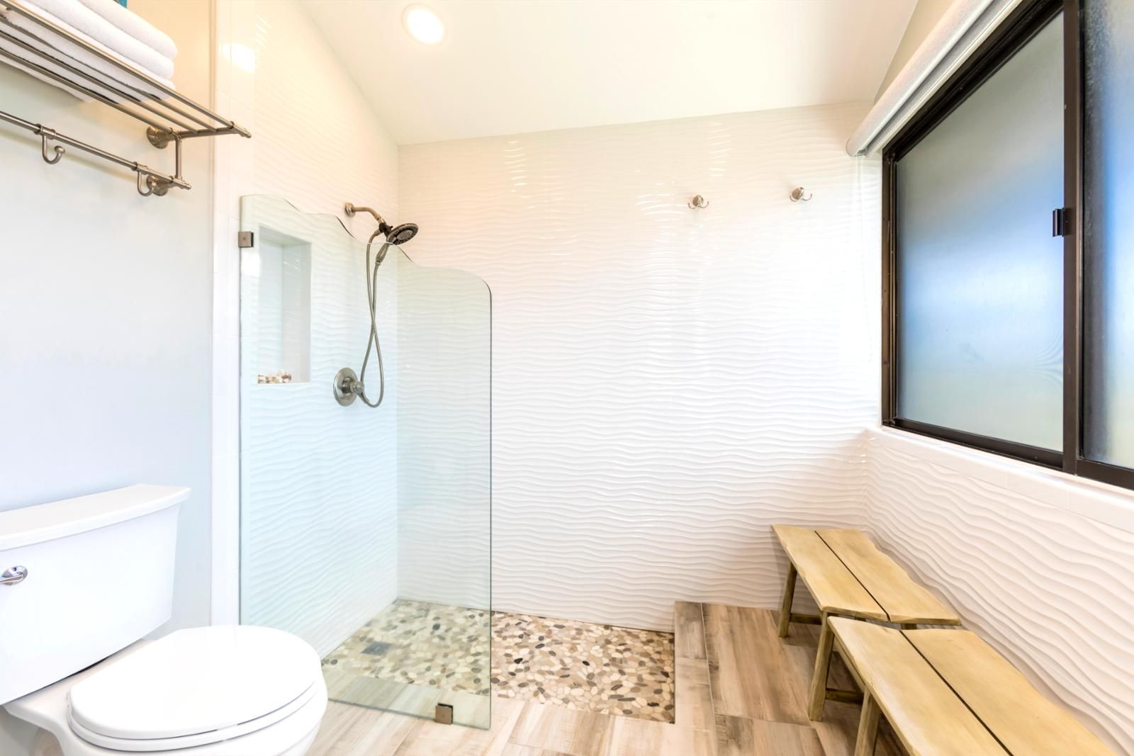 A bathroom with a glass shower enclosure, a toilet, a bench, and a window.