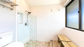 A bathroom with a glass shower enclosure, a toilet, a bench, and a window.