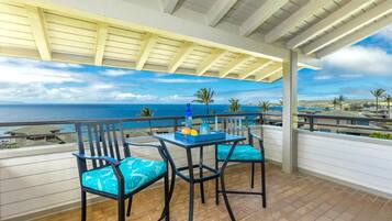 A balcony with a view of the ocean, two chairs, a table with a bottle and fruit, and a pergola overhead.