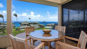 A covered outdoor dining area with a glass table and wooden chairs, overlooking a resort with multiple buildings and a view of the ocean. - Kapalua Bay Villas | 3 Oceanfront Villas, Sleeps 16 | Car Incl. w/ 6+ Nights | ML-4709 by KBM (Lahaina)