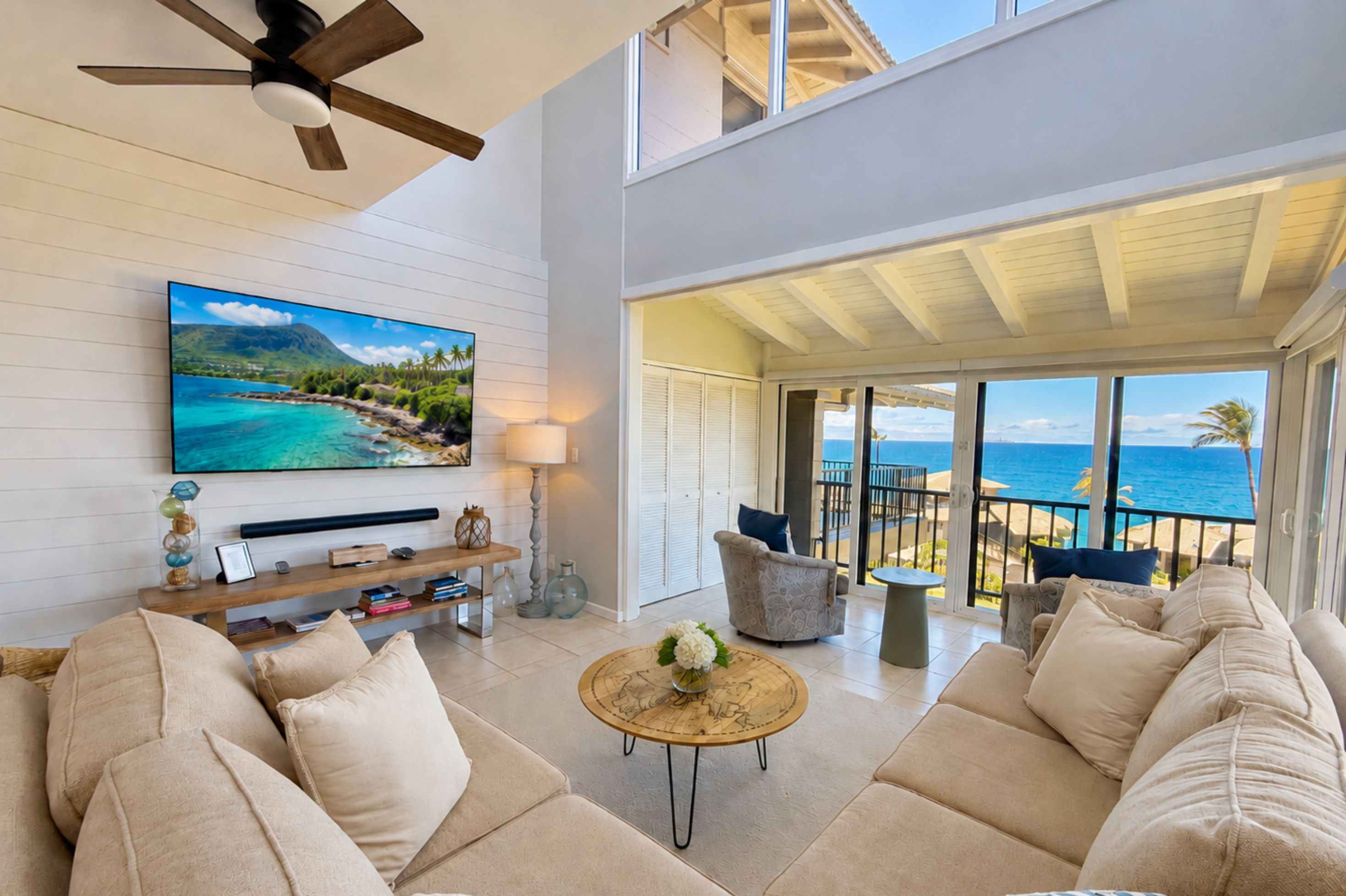 A spacious living area with a large flat-screen TV, comfortable seating, a ceiling fan, and a view of the ocean through large windows.