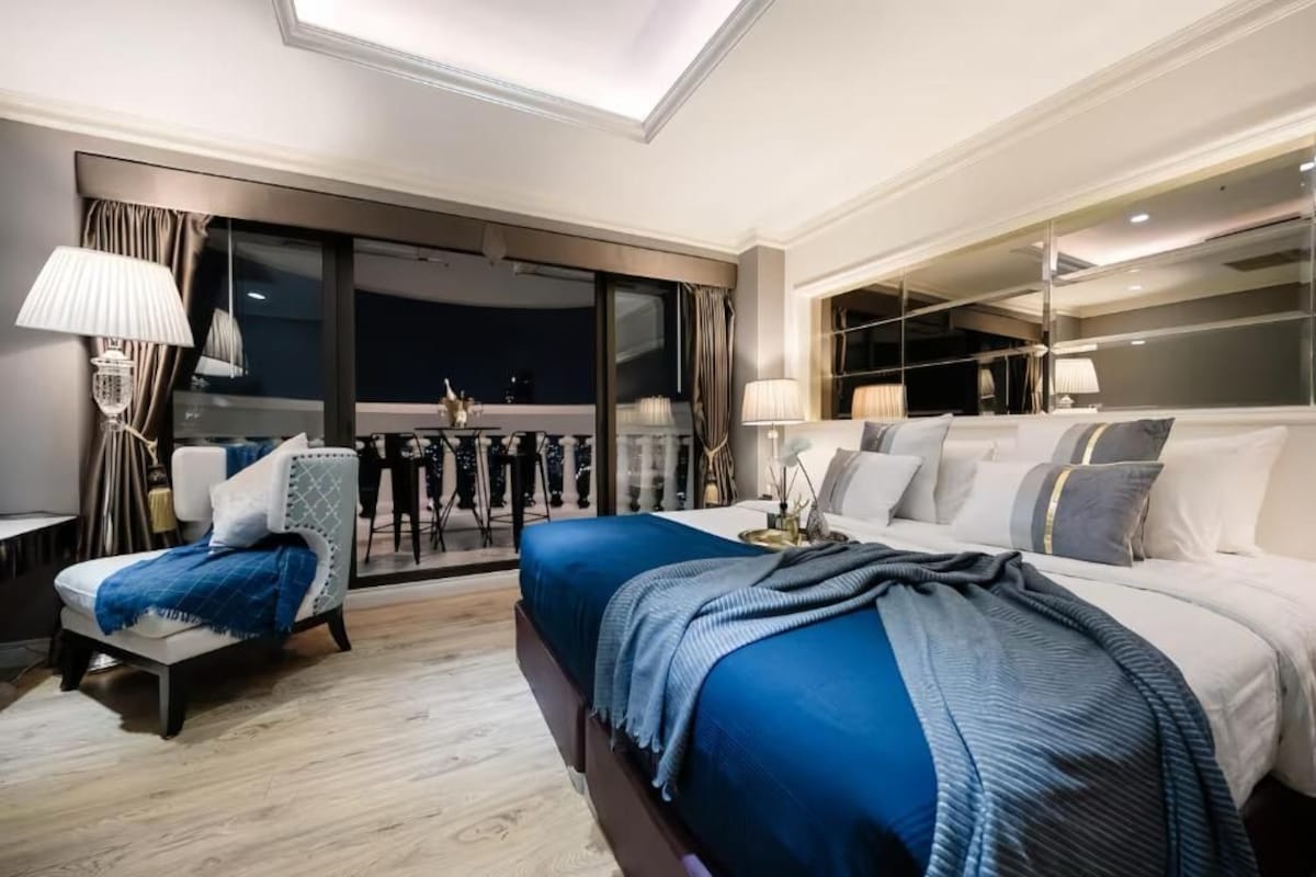 Habitación de hotel moderna con una cama grande, una zona de estar con una silla y una mesa, y un balcón con vistas.