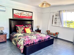 A bedroom with a bed, a nightstand, a chair, a window with curtains, and a painting on the wall. - Villa Anj'Any, 3 bedrooms, swimming pool, 300m from Grande Anse (Deshaies)
