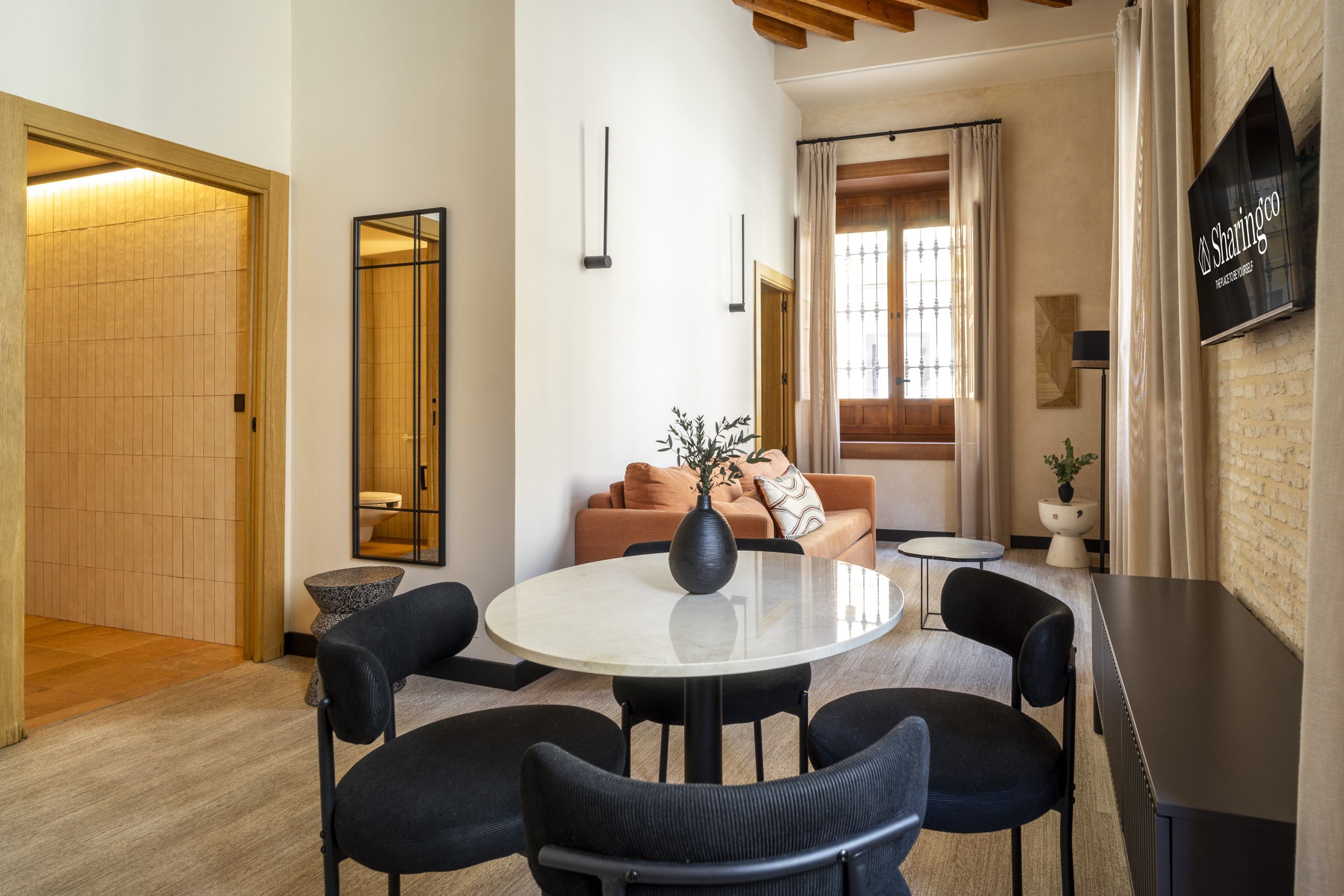 Un interior moderno con una mesa de comedor redonda, sillas negras, un sofá con cojines, una planta en maceta y un televisor fijado a la pared.