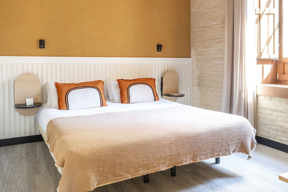 Un dormitorio con techo de madera, una cama con ropa de cama blanca y naranja, una mesita de noche y una ventana con cortinas.