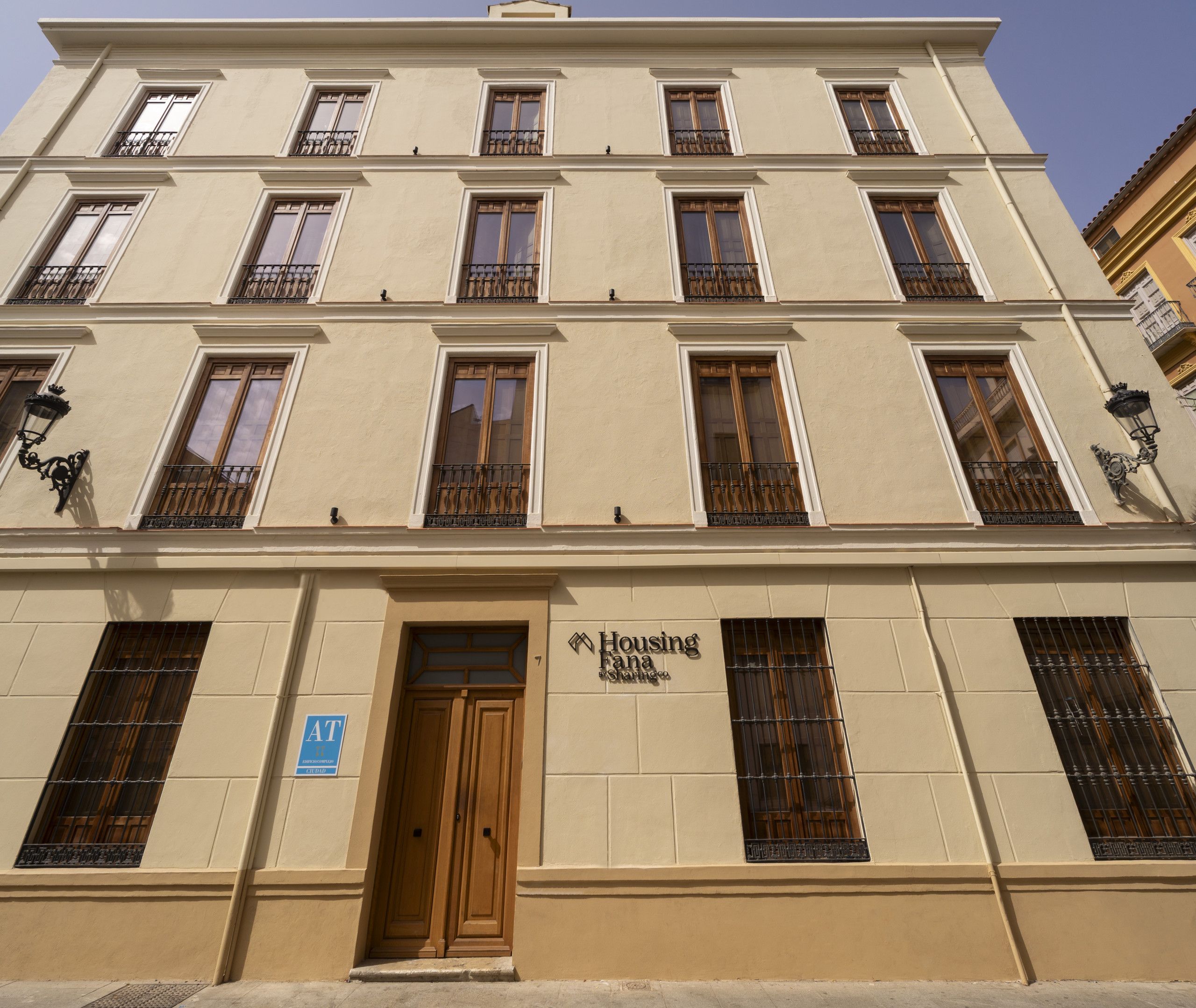 A multi-story building with a beige facade, multiple windows with wooden frames, and a central entrance with a wooden door.