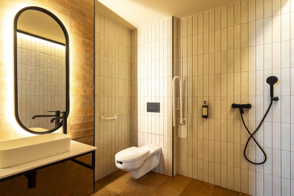 Un baño moderno con un espejo ovalado grande, un lavamanos blanco, un inodoro y una ducha con cabezal de ducha manual.