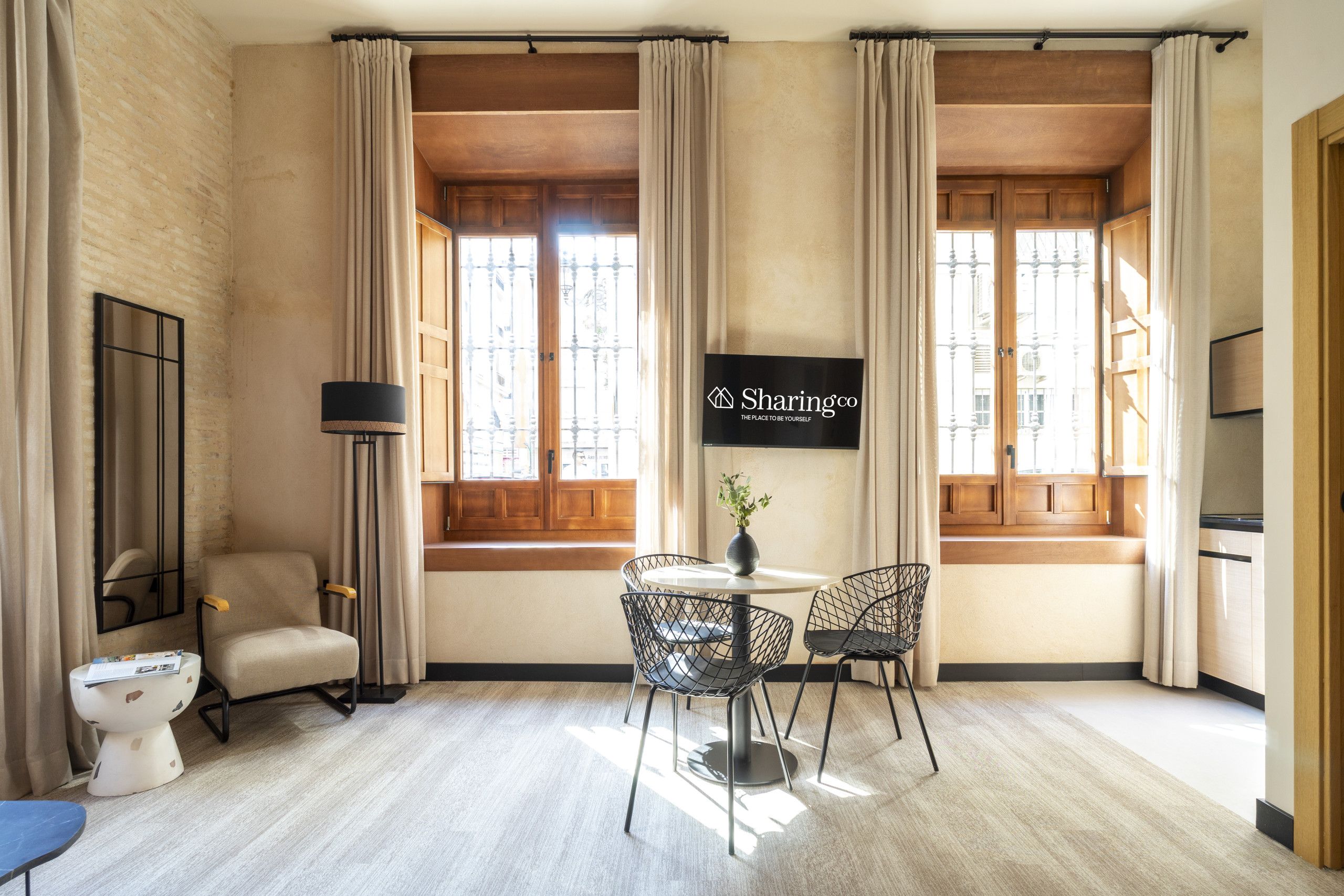 Habitación de hotel con un ventanal grande, un televisor de pantalla plana montado en la pared, una mesa redonda con dos sillas, una lámpara de pie y un sofá.