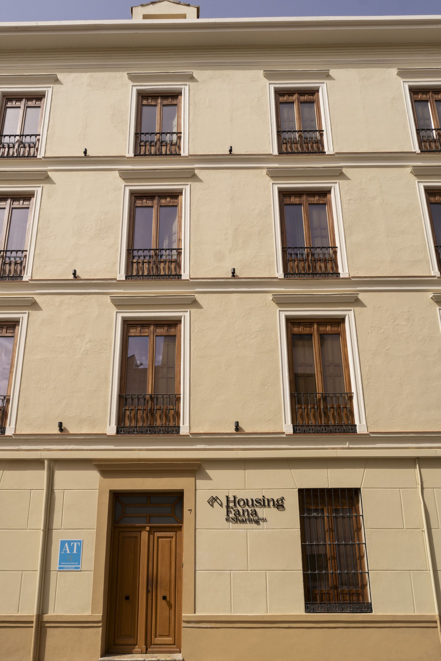 Un edificio de varios pisos con un cartel que dice 'Housing Fana Sharing Co.' y un letrero azul que indica 'AT' para acceso.