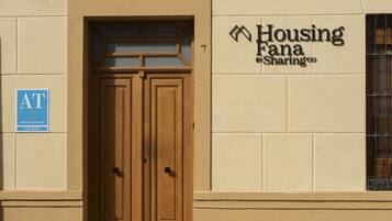 Entrada de un edificio con un par de puertas de madera, un letrero que dice 'Housing Fana Sharingco' y un cartel azul con 'AT CIUDAD' a la izquierda.