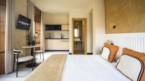 Quarto de hotel com cama, escrivaninha com cadeiras, televisão e vista para a cozinha.