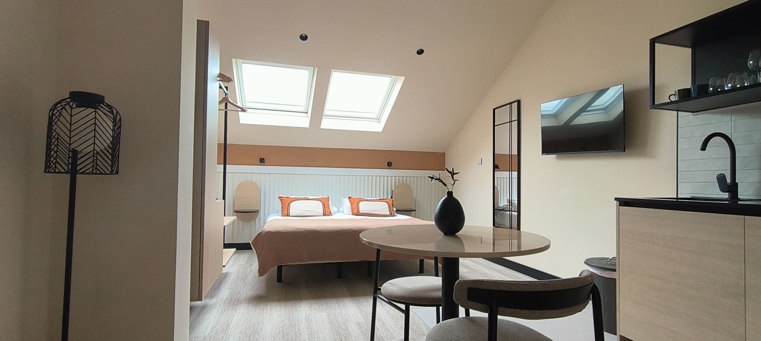 A modern hotel room with a bed, a round dining table, a small kitchen area, and a TV mounted on the wall.