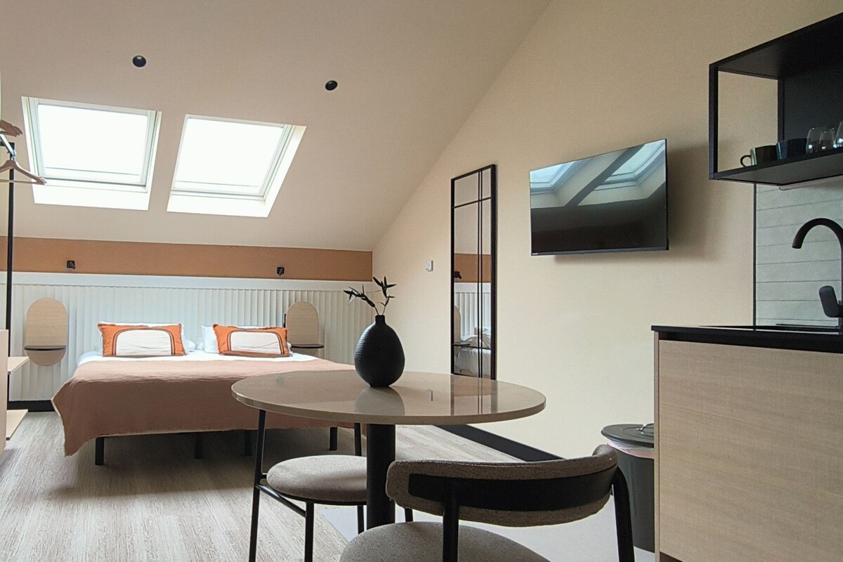 Una habitación de hotel moderna con una cama, una mesa redonda de comedor, una pequeña cocina y un televisor montado en la pared.
