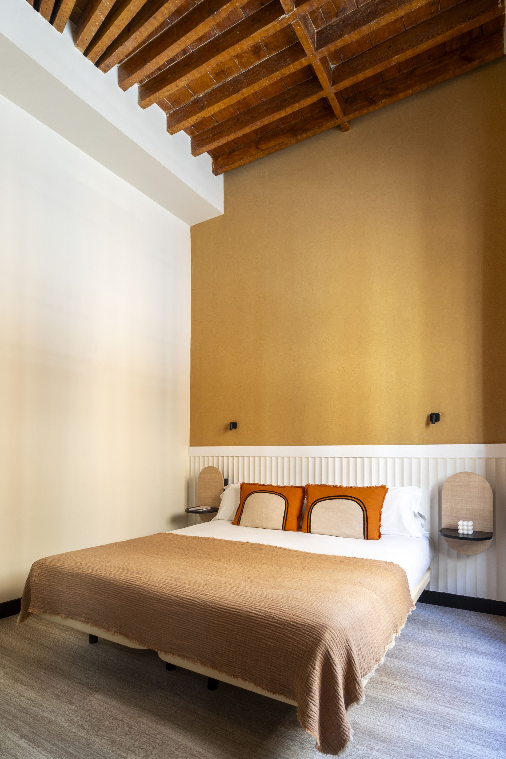 Un dormitorio con techo de madera, una cama con manta color beige y cabecera blanca.