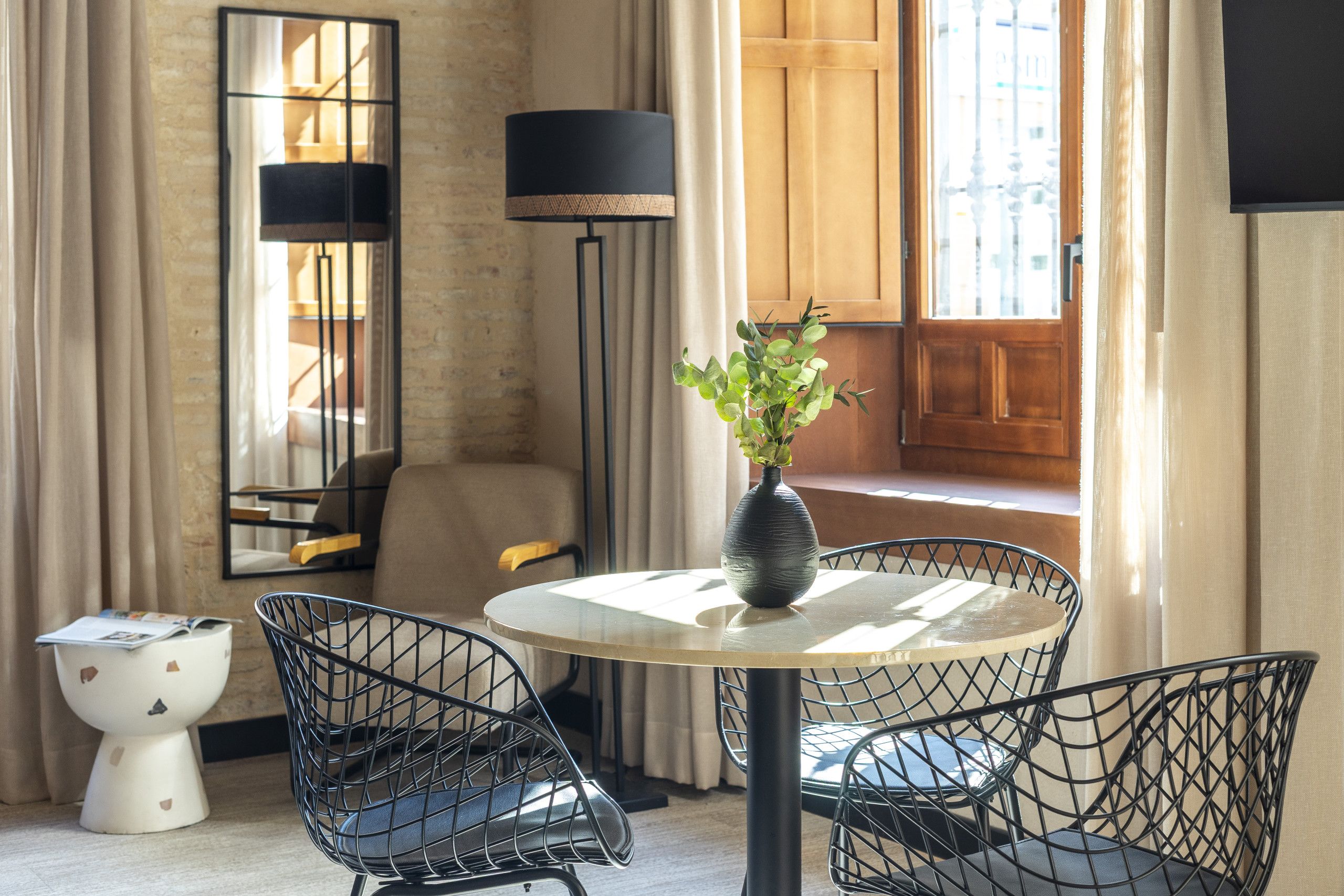 Habitación moderna con una mesa redonda, dos sillas de estructura metálica, un florero con follaje verde, una lámpara de pie y un espejo grande.