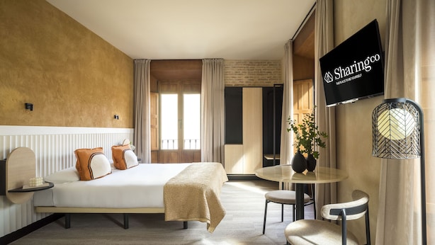 Quarto de hotel com cama, mesas de cabeceira, mesa redonda com cadeiras, TV fixada na parede e janela com cortinas.