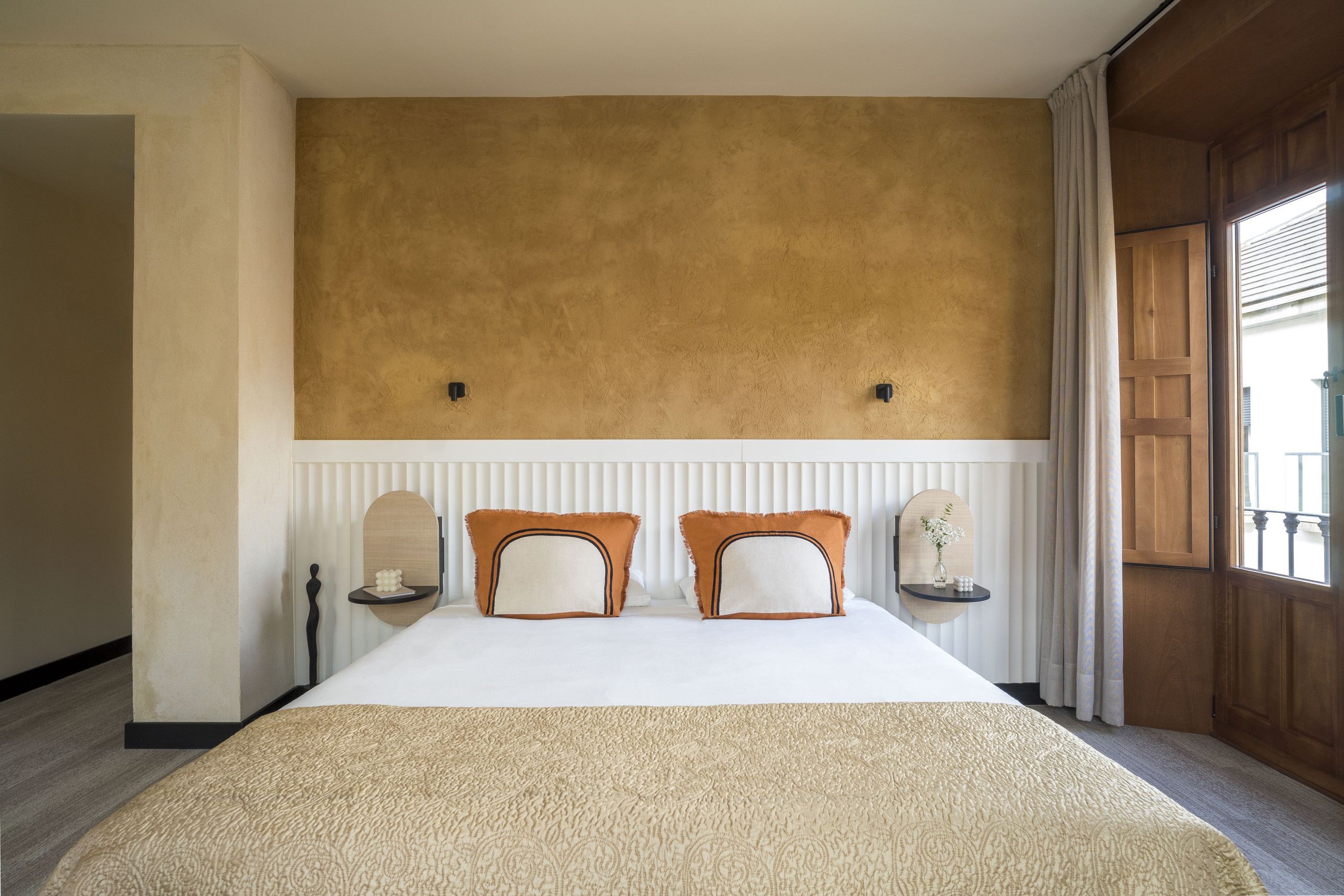 Una habitación de hotel con una cama grande, dos mesitas de noche, un estante en la pared y una ventana con cortinas.