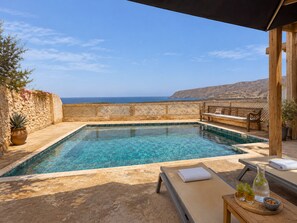 Una piscina rectangular con agua azul cristalina, rodeada por una cubierta de madera y un muro de piedra.