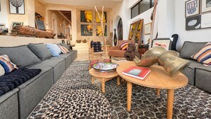Ein großzügiger Wohnraum mit hohen Decken, einem großen Fenster und Blick ins Innere. Es gibt eine Sofagarnitur, einen Couchtisch und einen gemusterten Teppich.