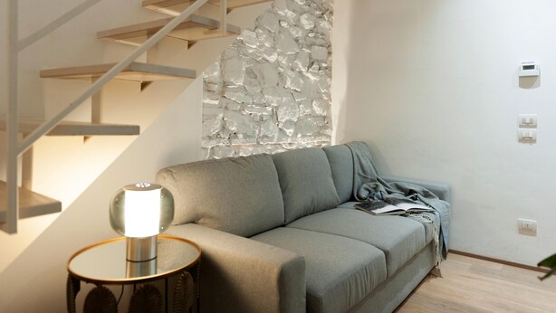 Ein modernes Wohnzimmer mit einem grauen Sofa, einem runden Beistelltisch mit einer Lampe, einer Holztreppe und einer strukturierten Wand.