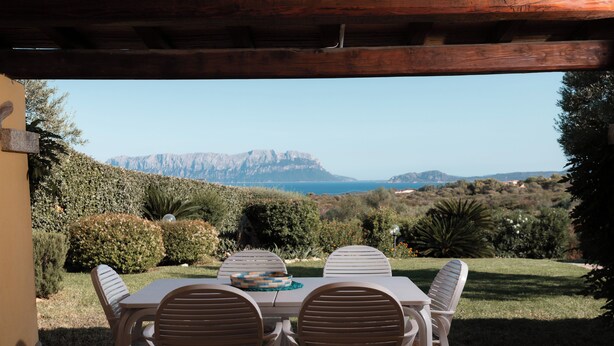 Um pátio ao ar livre com mesa e cadeiras, com vista para uma paisagem montanhosa e o mar.
