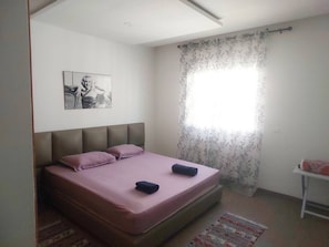 Ein Schlafzimmer mit Bett, einem Kopfteil, einem Fenster mit Vorhängen, einem kleinen Tisch und einem Teppich.