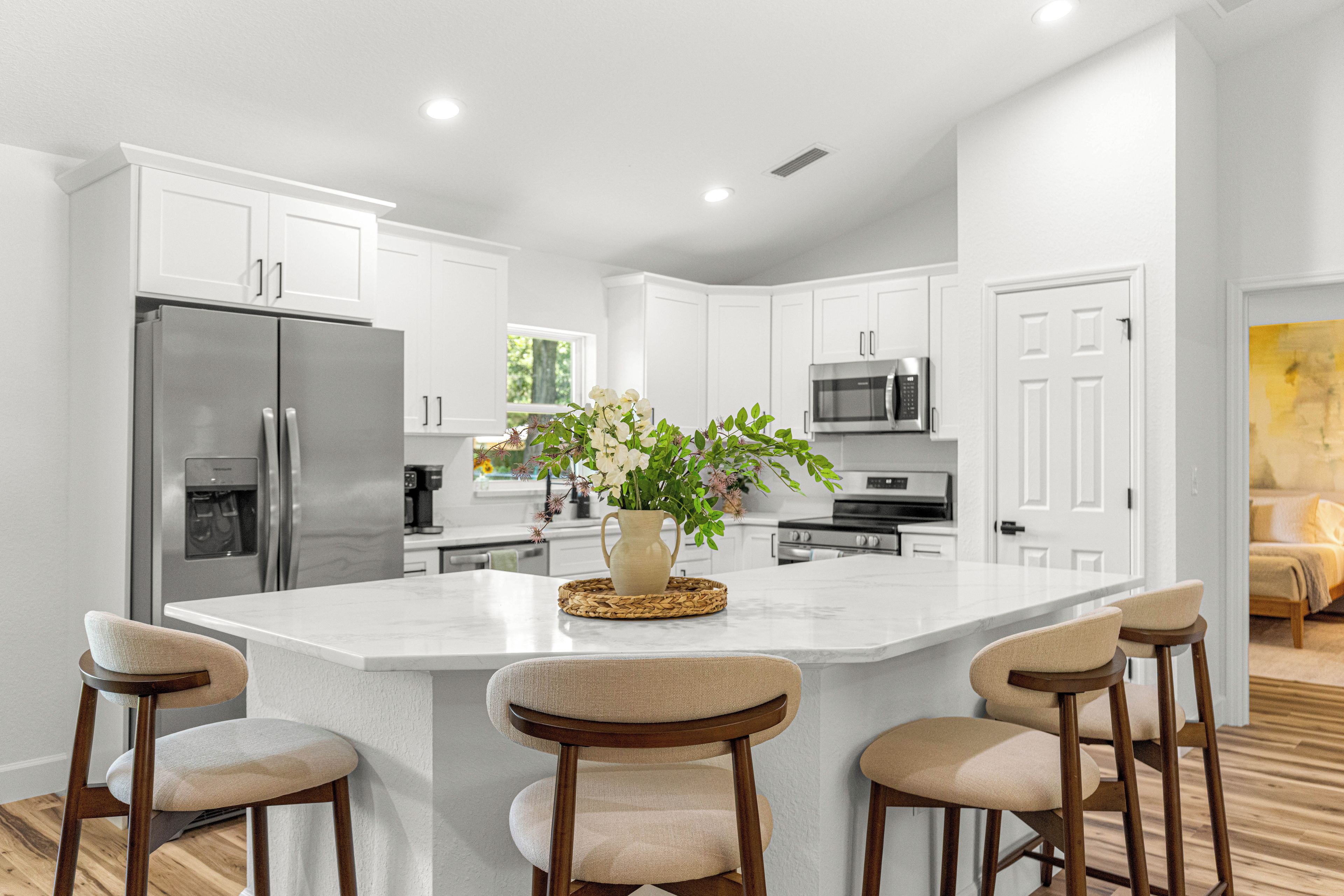 A modern kitchen with white cabinets, a central island with bar stools, a refrigerator, a microwave, and a coffee machine.