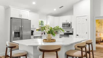 A modern kitchen with white cabinets, a central island with bar stools, a refrigerator, a microwave, and a coffee machine.