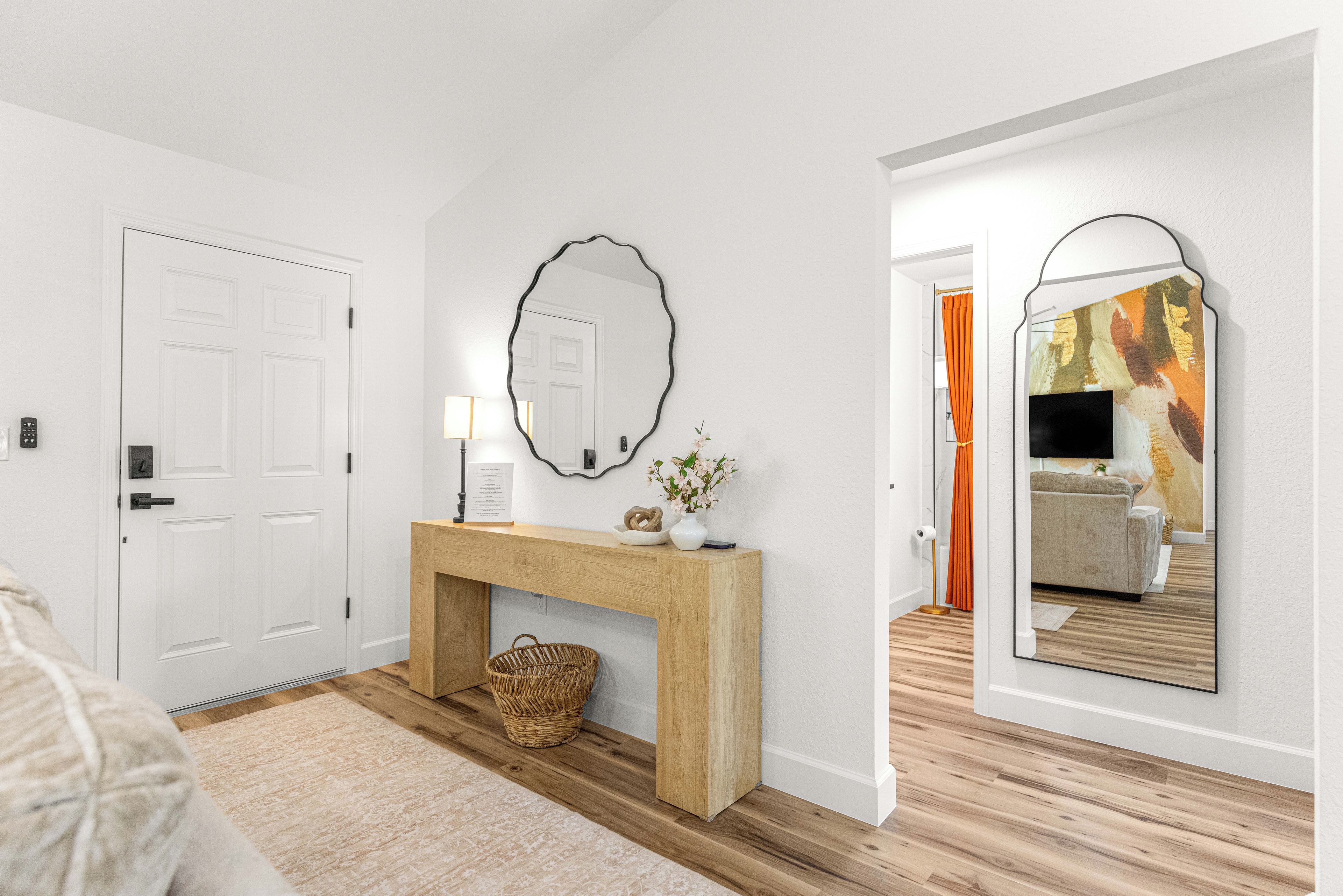 A hallway with a white door, a wooden console table with a mirror, a wicker basket, and a framed picture on the wall.