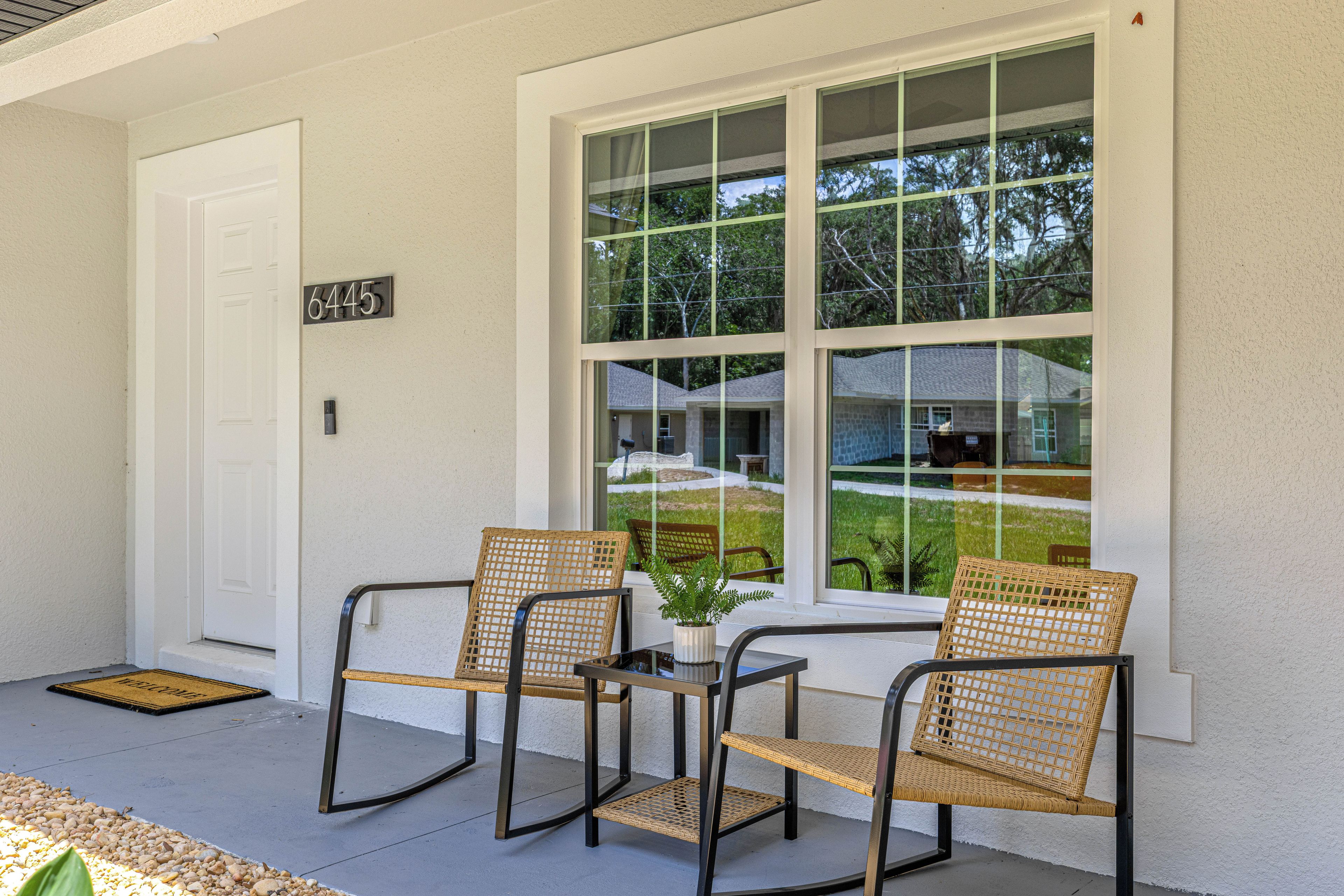 A covered porch with two wicker chairs, a small table, and a window with a view of a house and trees.