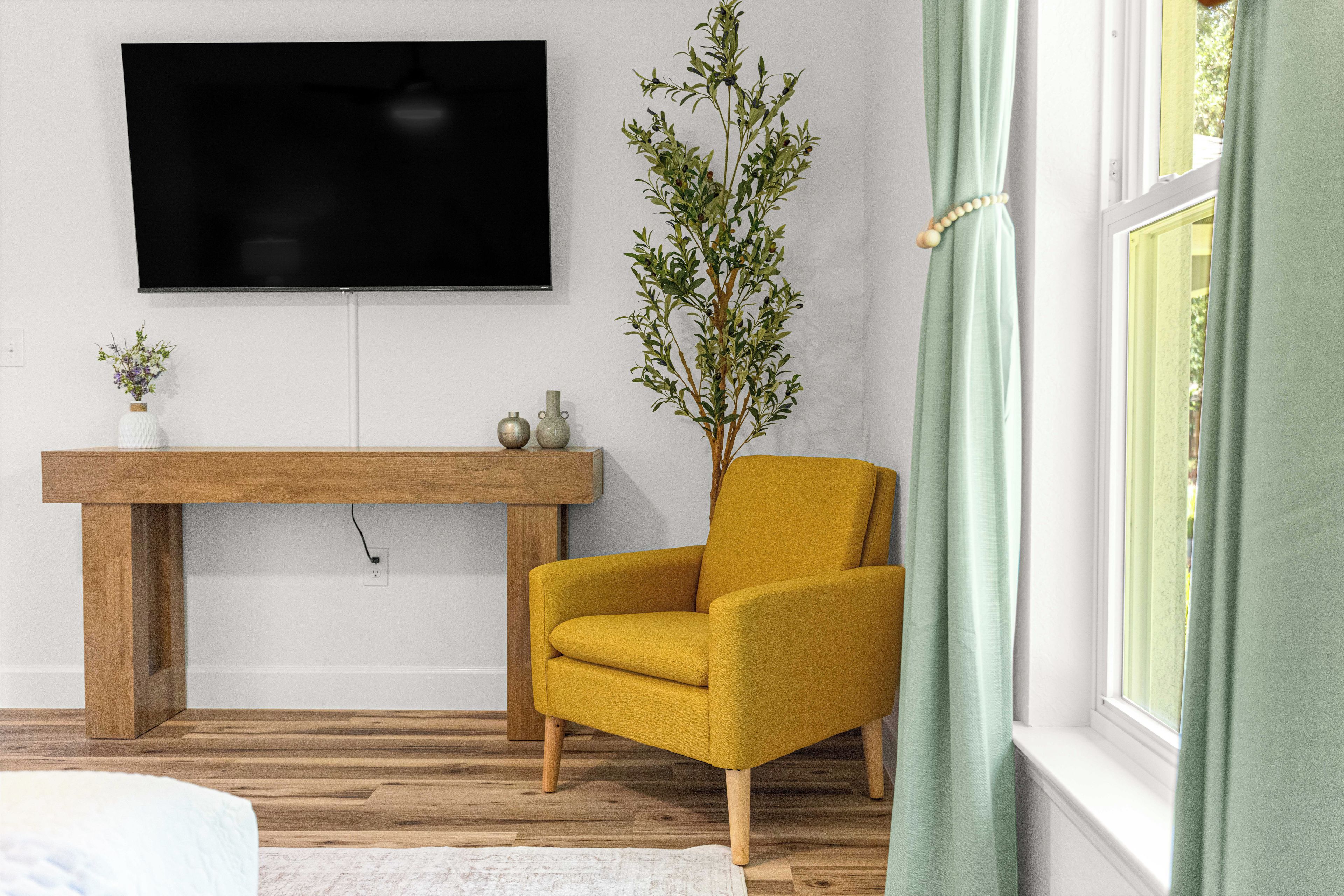 En stue med en gul lænestol, et træbord, et fladskærms-tv og en potteplante.