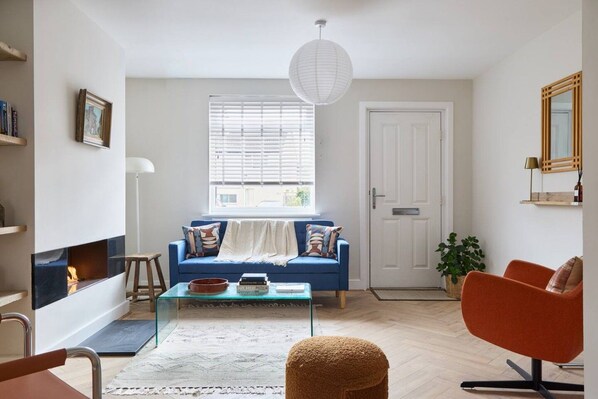 A living room with a blue sofa, a glass coffee table, a fireplace, a door, and a large window with blinds.