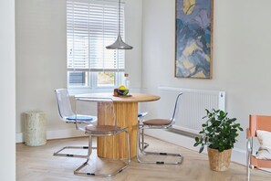 A modern dining area with a round table, two chairs, a hanging light, and a painting on the wall.