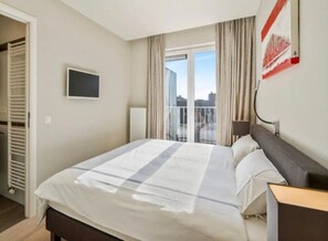 A hotel room with a bed, a television, a window with curtains, and a view of the city. - Apartment in Blankenberge With Ocean Views (Blankenberge)