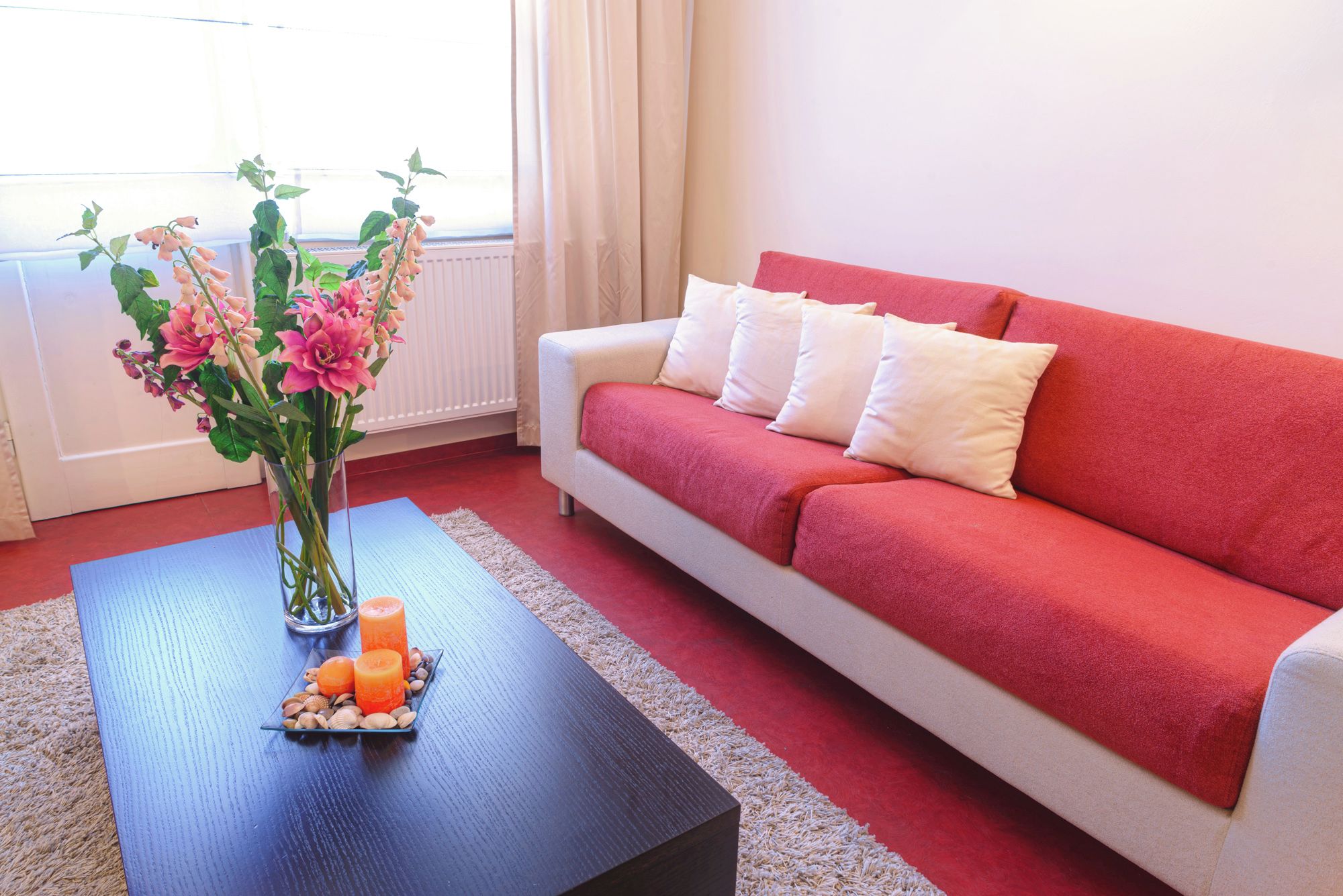 A room with a red sofa, a glass vase with flowers, and a dark wooden table with candles and decorative stones.